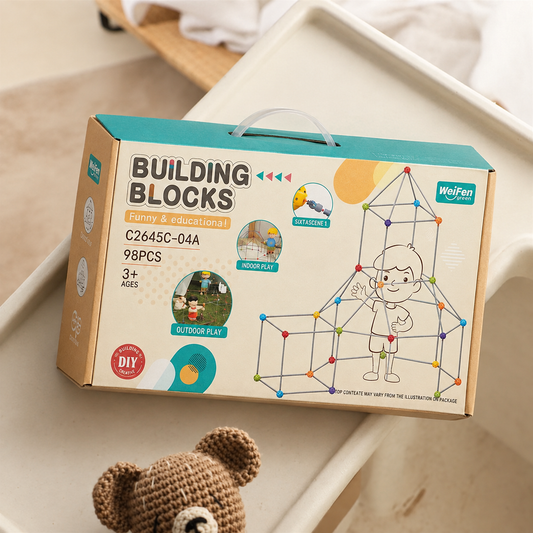 A boxed children's building toy set labeled 'BUILDING BLOCKS' sits on a light beige tray. The package is tan cardboard with teal and yellow accents, printed graphics of a child assembling geometric blocks, and small circular callouts including 'outdoor play.