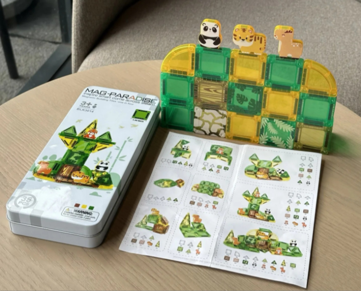MAG PARADISE magnetic building toy set displayed on a light wood table, showing the illustrated metal tin box, a printed instruction sheet, and a green/yellow translucent magnetic block tower with animal-shaped tiles on top.