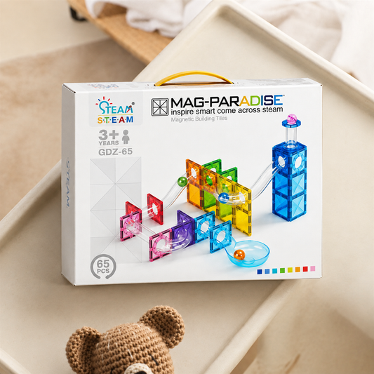 A boxed children's magnetic building tile set labeled MAG-PARADISE STEAM, shown standing on a beige tray with a small teddy bear visible at the bottom. The box is white with colorful printed tile examples and age/piece information.