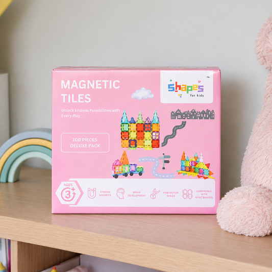 A pink box of Shapes for Kids Magnetic Tiles placed on a wooden shelf next to a plush toy, with a rainbow toy visible in the background.