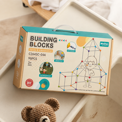 A boxed children's building toy set labeled 'BUILDING BLOCKS' sits on a light beige tray. The package is tan cardboard with teal and yellow accents, printed graphics of a child assembling geometric blocks, and small circular callouts including 'outdoor play.