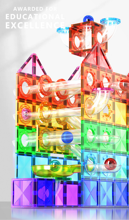 A colorful magnetic tile play set constructed into a complex marble run tower with transparent acrylic blocks in red, orange, yellow, green, blue, and purple, with small balls inside curved tubes. The image has a white background with a faint reflection and the text 'AWARDED FOR EDUCATIONAL EXCELLENCE' in the top left corner.